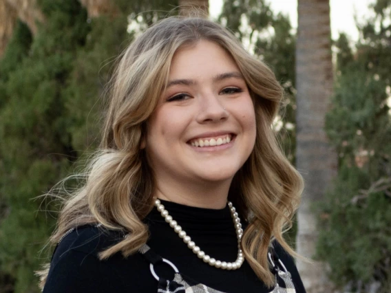 Photo of woman with blonde shoulder length hair, wearing a pearl necklace and a black shirt with black and white patterned overalls, she is smiling at the camera