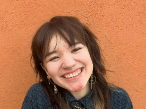 Ree Lee, a young person with shoulder-length brown hair, wearing a blue top, standing in front of an adobe wall and smiling