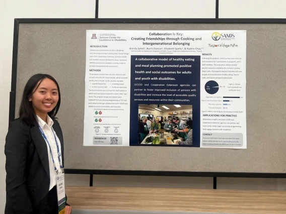 Nadine Chau, an Asian individual with shoulder-length black hair, is standing next to their poster. They are wearing a white collared button-up shirt, a black blazer, and an AUCD lanyard.