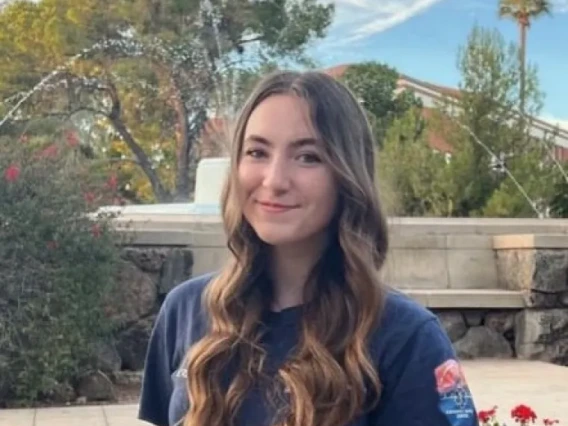 Ella Randolph, a young woman with long brown hair, wearing a dark blue t-shirt, standing outside and smiling