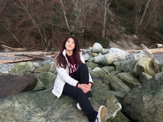 Anahy Fernandez sitting on a large rock smiling. Anahy has long red-brown hair, wearing winter wear, and is holding on to one leg while the other is extended.