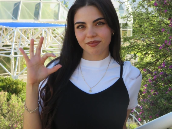Subject is in the center of the picture, smiling and holding up her right arm, doing the "Wildcat" symbol (pinkie, ring and middle finger spread open, pointer and thumb curved into a "C" shape). Subject is wearing a plain white short sleeve, with a black jumpsuit over it. Subject is standing in front of Biosphere 2.