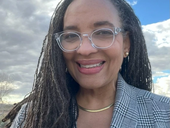 Woman wearing a blue plaid blazer and pink shirt, she has long box braids wearing clear rimmed glasses