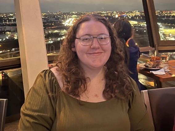 A woman sitting in front of a skyline view wearing a dark green shirt. She has mid length curly red hair and has brown glasses on. She sits at a dining table with a smile looking towards the camera.
