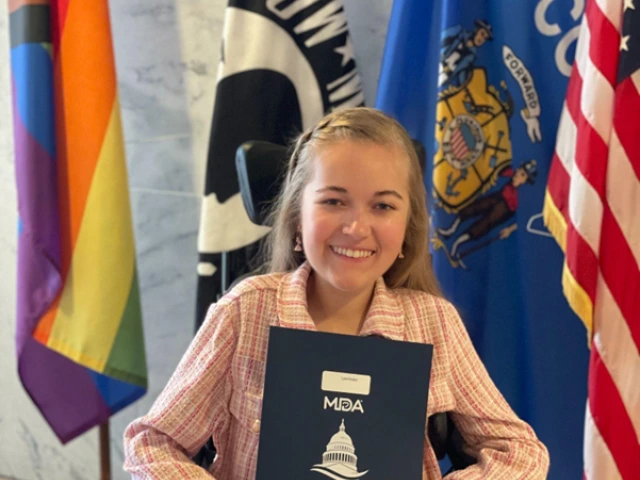 Reagan, a white woman in a pink suit and who sits in a power wheelchair, holds up a folder that says MDA Advocacy on the cover. Reagan sits in front of several inclusive flags, alongside the state of Wisconsin flag and the American flag. Reagan smiles at the camera.