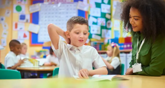 Elementary aged child sitting at work table reading a book with oversight from teacher