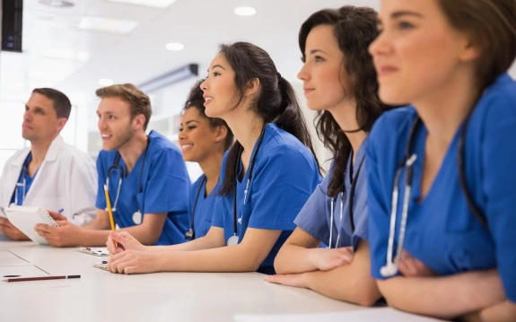 two young men and four young women sit at a table and look ahead. They are wearing blue scrubs with stethoscopes around their necks.