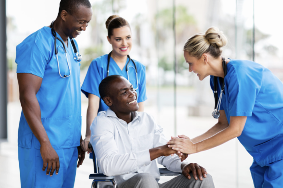 a young man sits in a wheelchair and is shaking hands with a young nurse with blond hair. A male nurse and another female nurse stand behind the wheelchair and smile