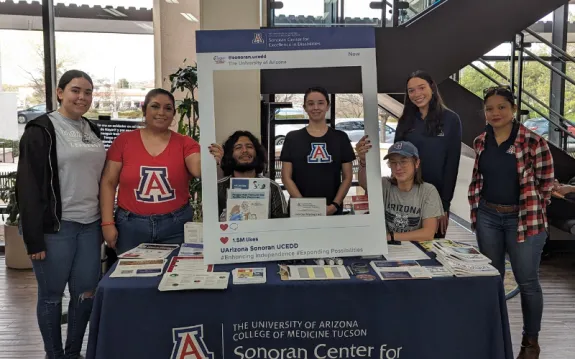 The author (third from right) sits with Sonoran Center staff at a conference table. They are holding a large poster board with a square cut in the middle, and decorated to resemble a social media post. The table has numerous Sonoran Center fliers and brochures on it.