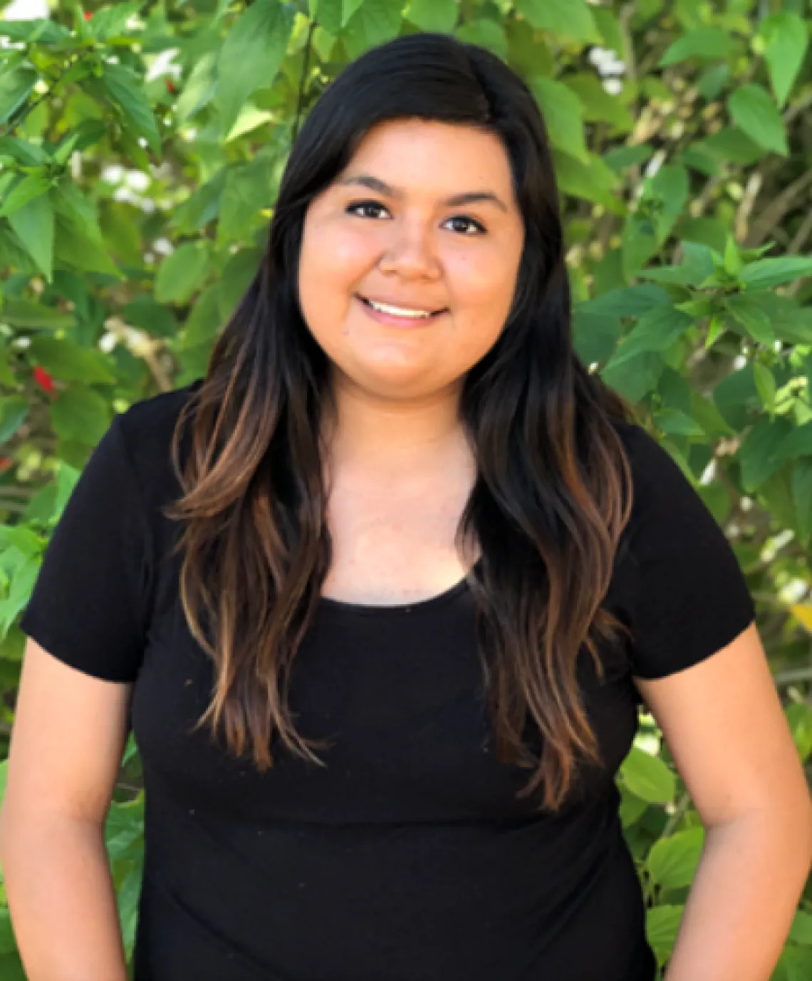 Photo of Bianca Aguilar, a woman with long brown hair wearing a black shirt and standing in front of greenery.