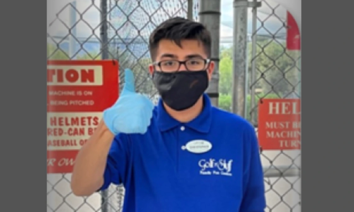 Young adult with short dark hair and glasses, wearing a blue shirt and a black medical mask, giving a thumbs-up to the camera.