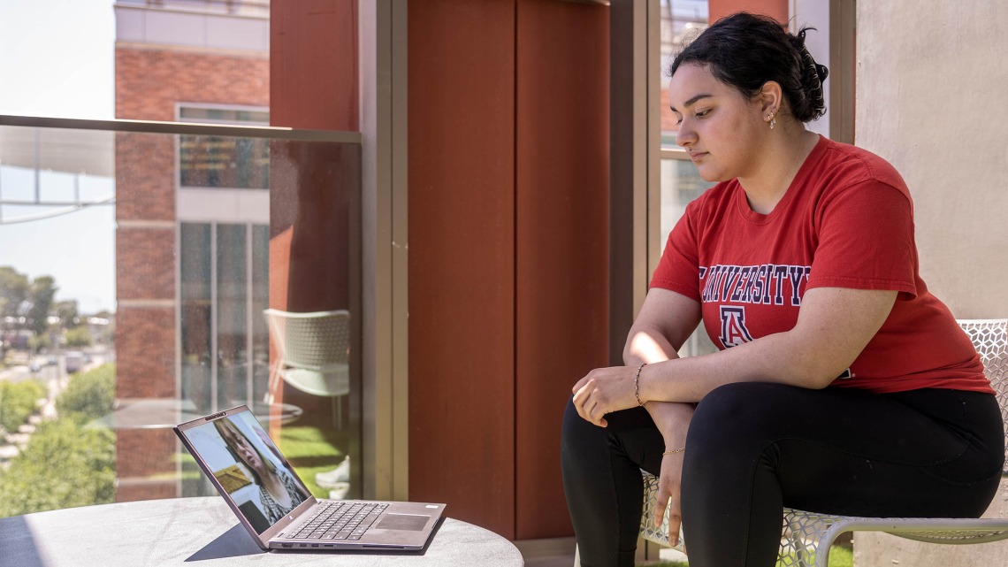 Student in a red University of Arizona shirt sitting with their hands crossed in front of them watching the interdisciplinary training session on their laptop.