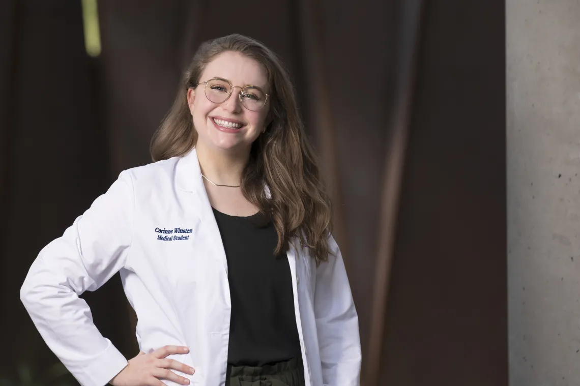 White woman with long light brown hair, wearing glasses and a white medical coat, smiling with her hand on her hip