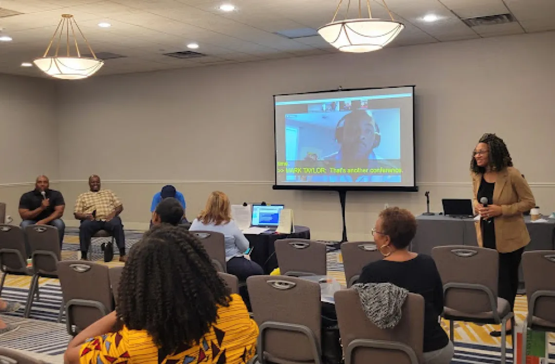 Crowded room of people watch a presentation being led by a Black woman with curly shoulder length hair, wearing a tan blazer