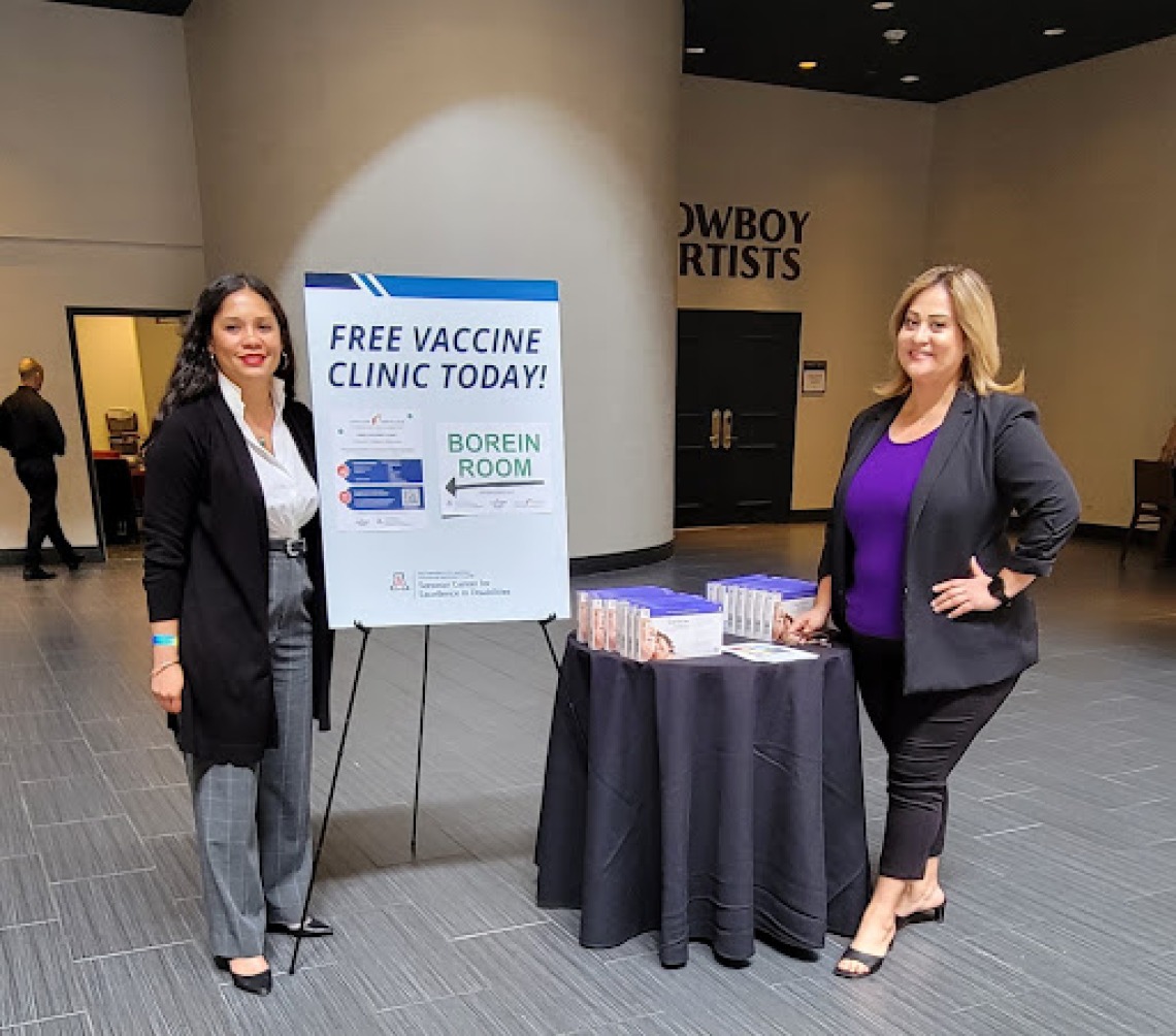 Two women pose on either side of a sign describing a free vaccine clinic