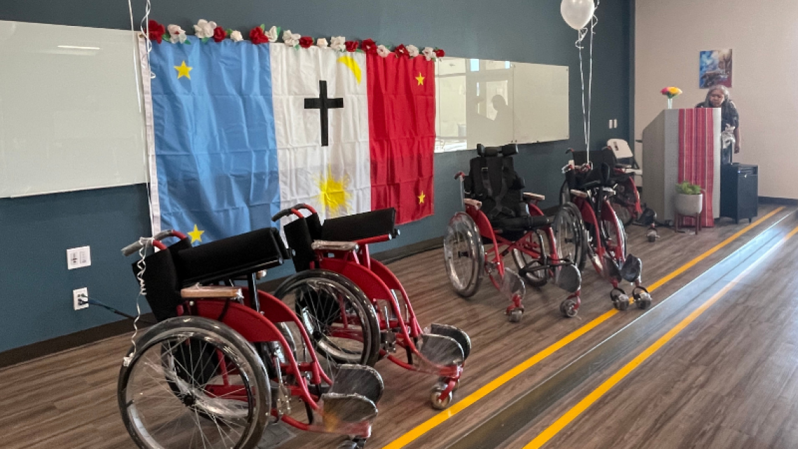 Several new red and black wheelchairs, still wrapped in cellophane, positioned side-by-side on a stage with a Tribal flag hung in the background.