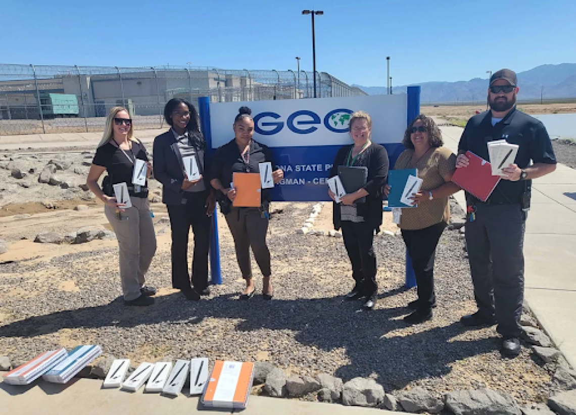 Six people holding LiveScribe pens and standing outdoors in front of a GeoGroup sign, with chain link fence and prison facility visible in the background