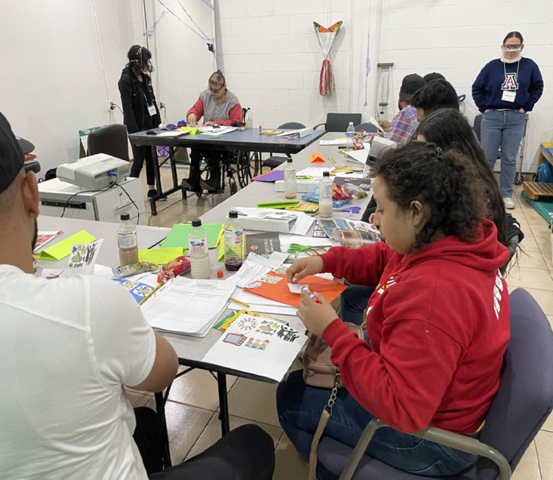 Participants in the DISCAPAZ scrapbooking workshop work on their creations, while Paulette Nevarez stands in the background, observing