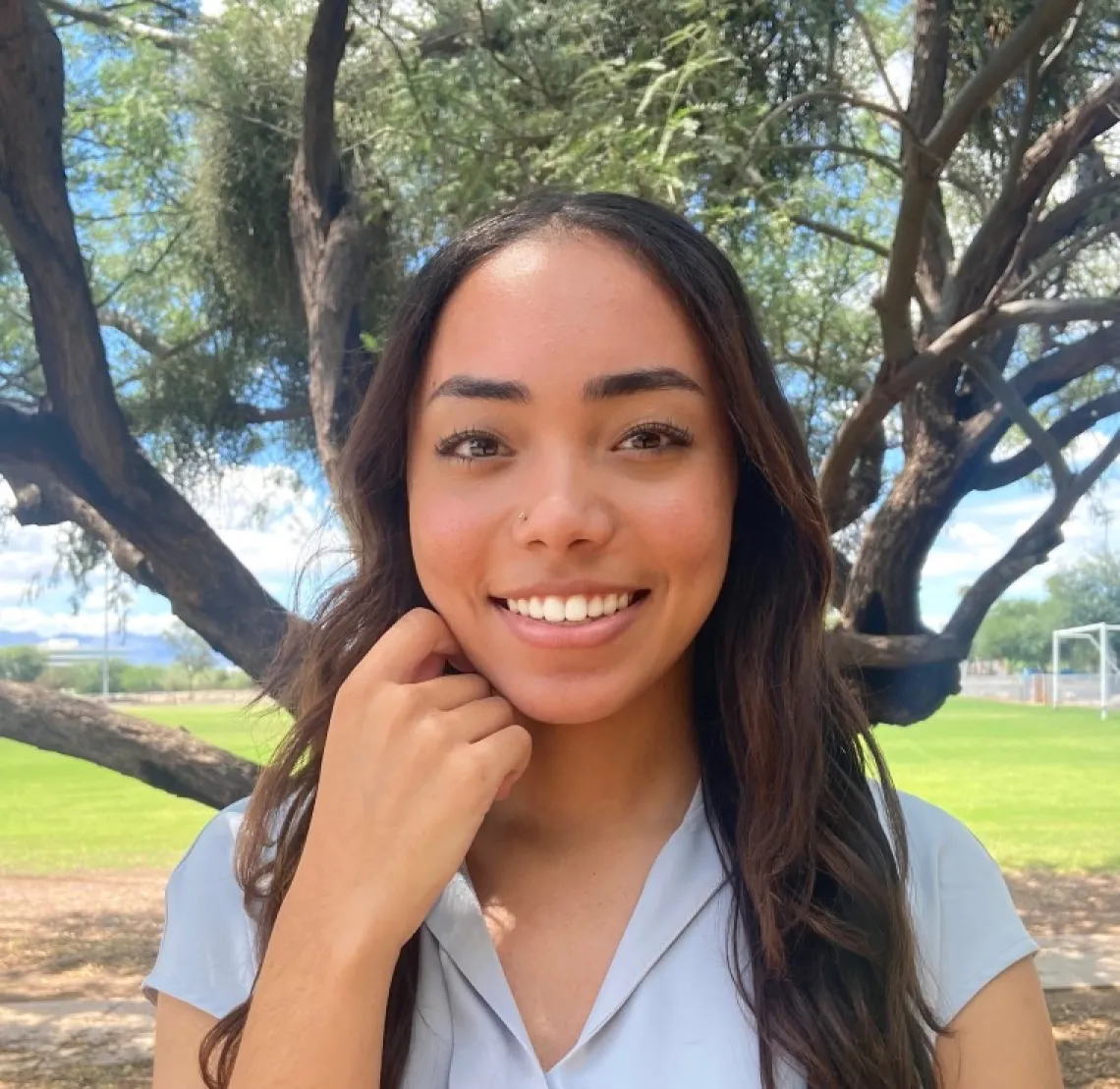 Aaliyah Washington, a young woman with long brown hair, wearing a light blue top, standing outside, in front of a tree, smiling