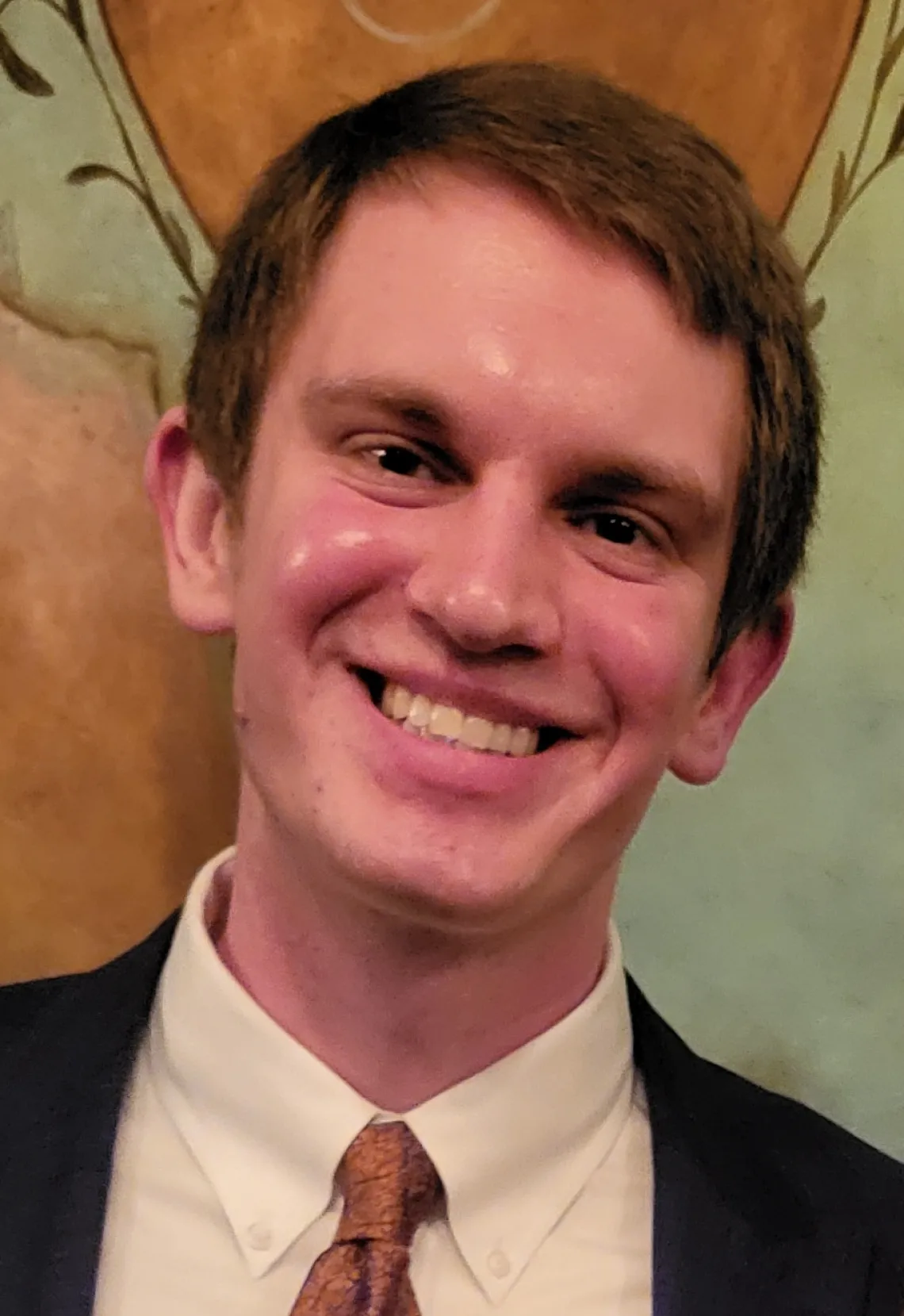 Drew Milne,  a white man in his late 20s with short brown hair, wearing a suit with a white shirt, copper tie, and blue jacket