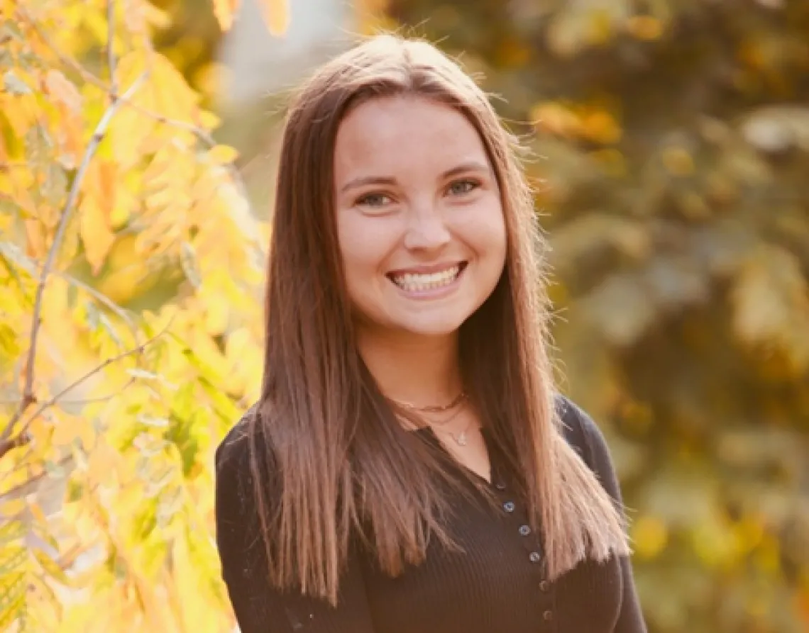 Samantha Zwillinger, a young woman with long brown hair, wearing a brown long sleeve top, standing outside among trees with yellow leaves, smiling