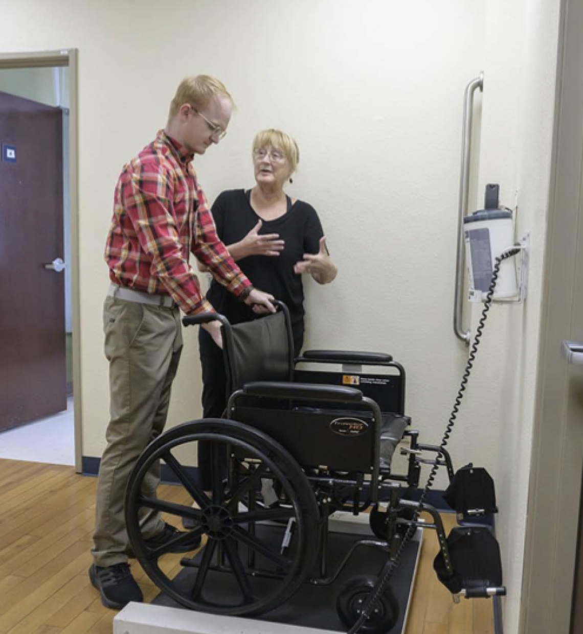 a gentleman is standing behind a wheelchair and holding onto its handles while a woman gestures with her hands beside him. The wheelchair is on a scale.