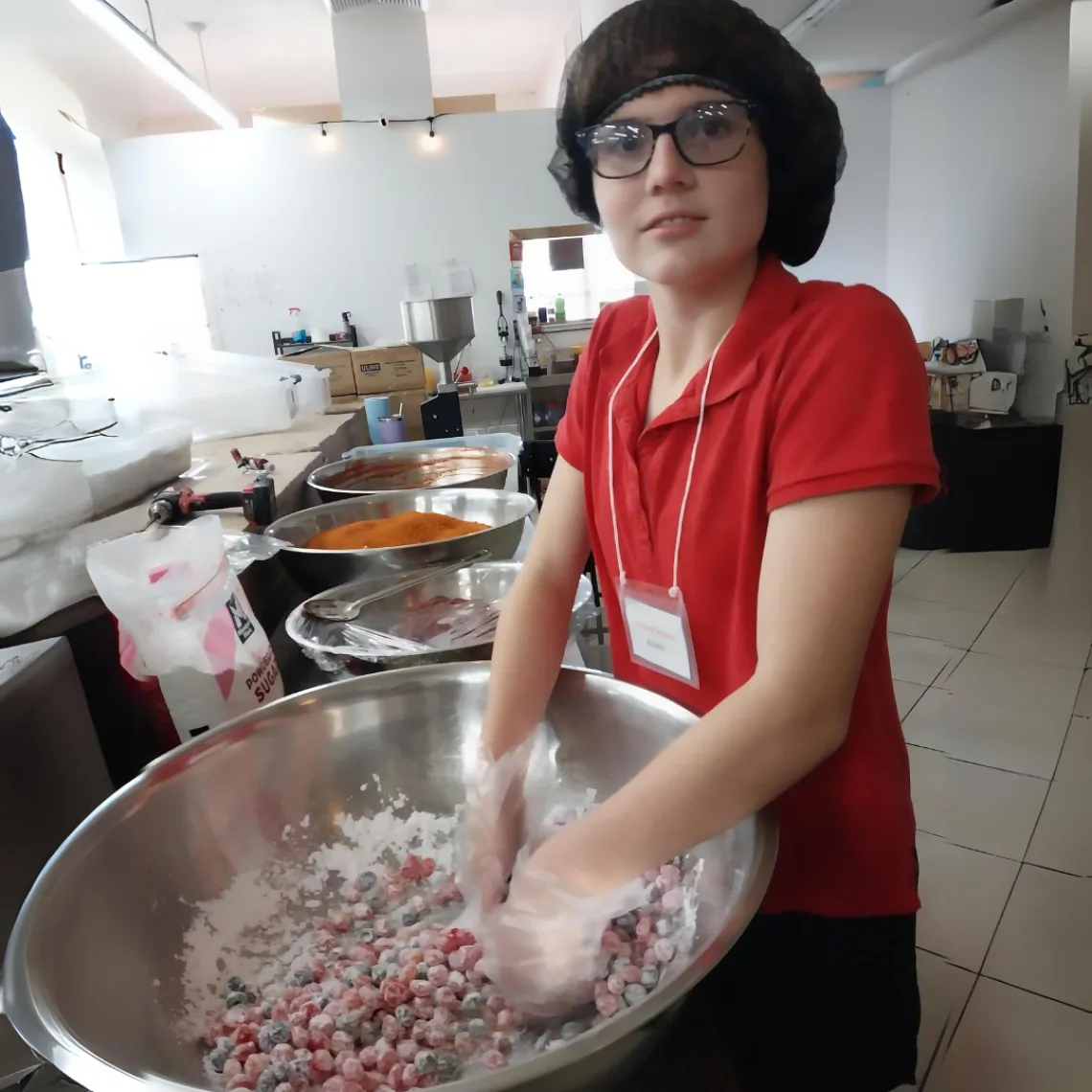 Student wearing a hairnet and gloves hand mixing food in a large commercial mixing bowl.