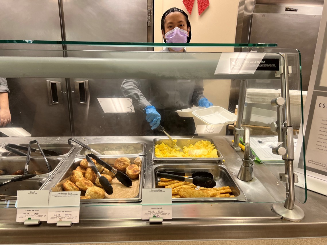 woman in hair net and surgical mask scoops scrambled eggs onto a plate behind a cafeteria counter 