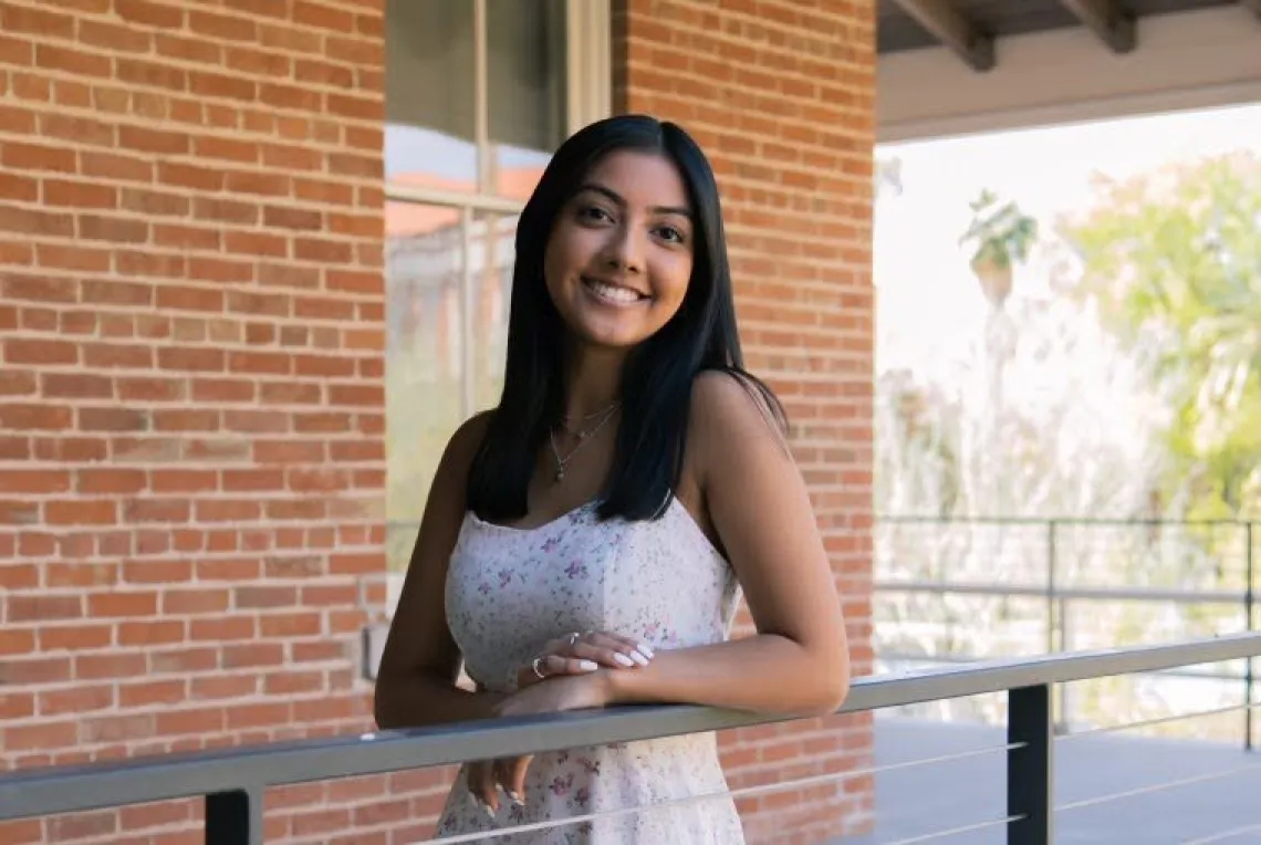 Victoria Almanza, a U of A student with long brown hair wearing a white top, standing outside in front of a brick building on a sunny day, smiling. 