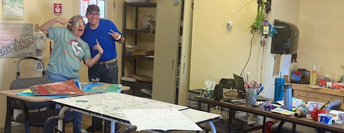 Two people posing and smiling in a classroom behind a desk covered in various artworks.