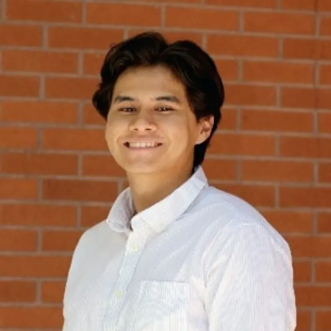 Joseph Fraire, a young person with dark hair wearing a white button-down shirt, standing in front of a brick wall and smiling