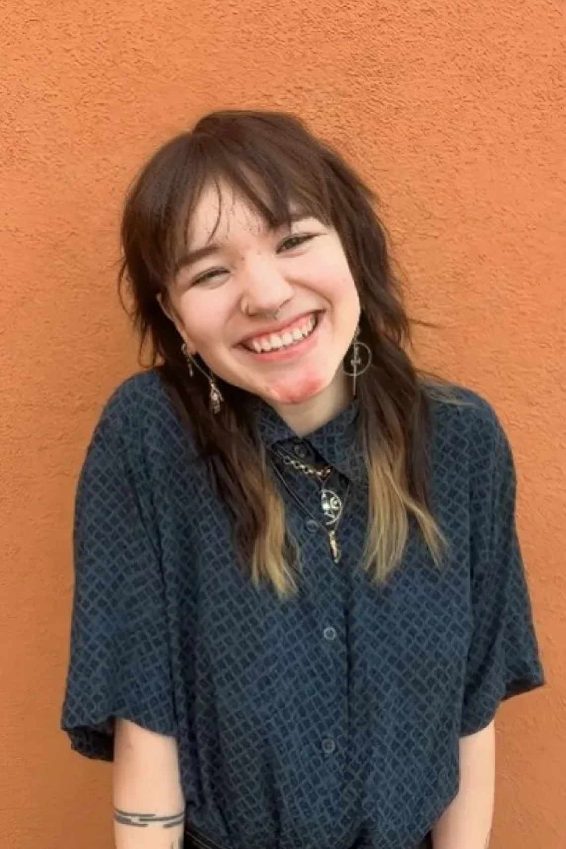 Ree Lee, a young person with shoulder-length brown hair, wearing a blue top, standing in front of an adobe wall and smiling