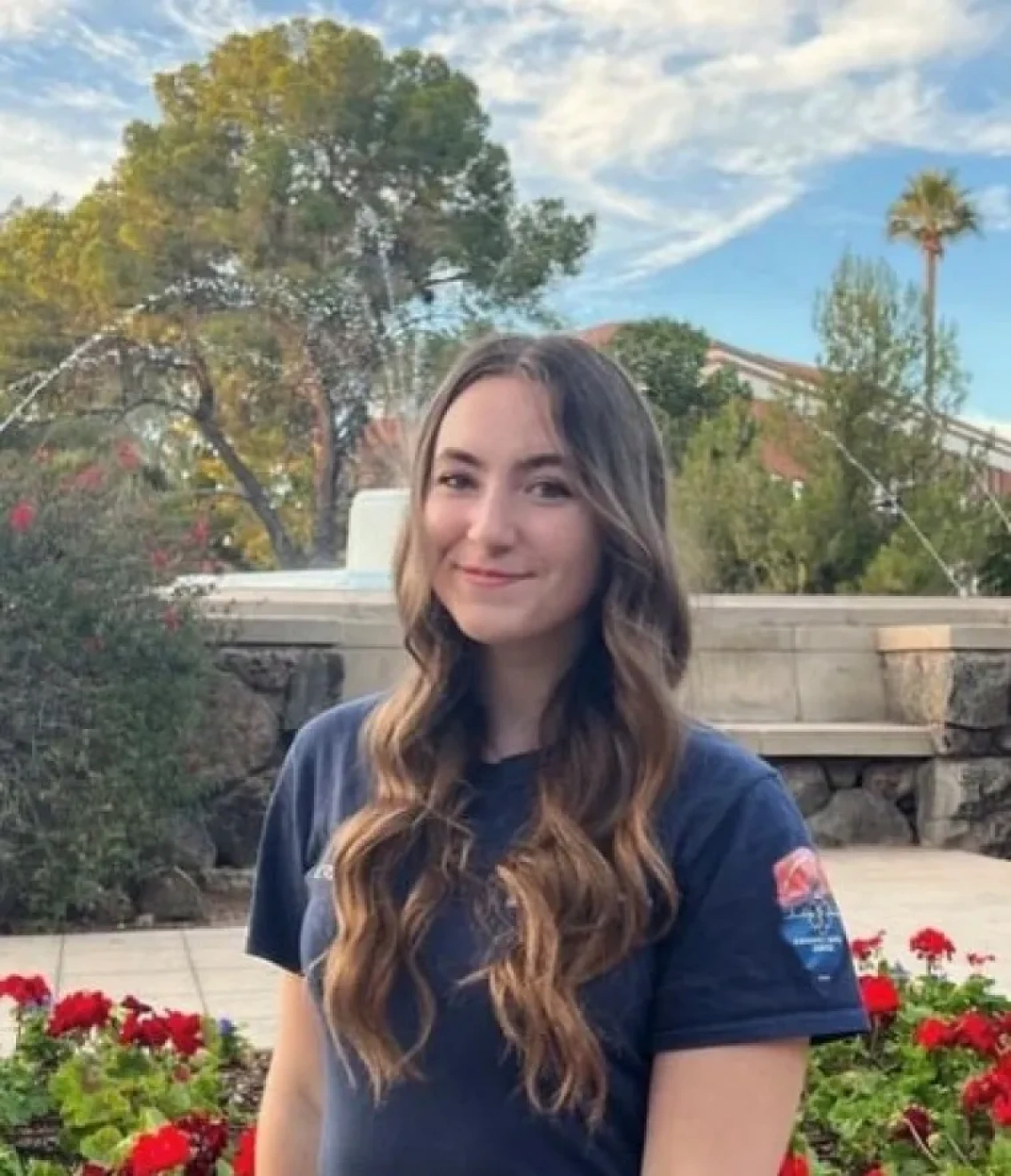 Ella Randolph, a young woman with long brown hair, wearing a dark blue t-shirt, standing outside and smiling