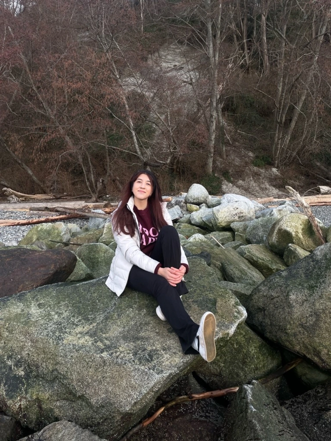 Anahy Fernandez sitting on a large rock smiling. Anahy has long red-brown hair, wearing winter wear, and is holding on to one leg while the other is extended.