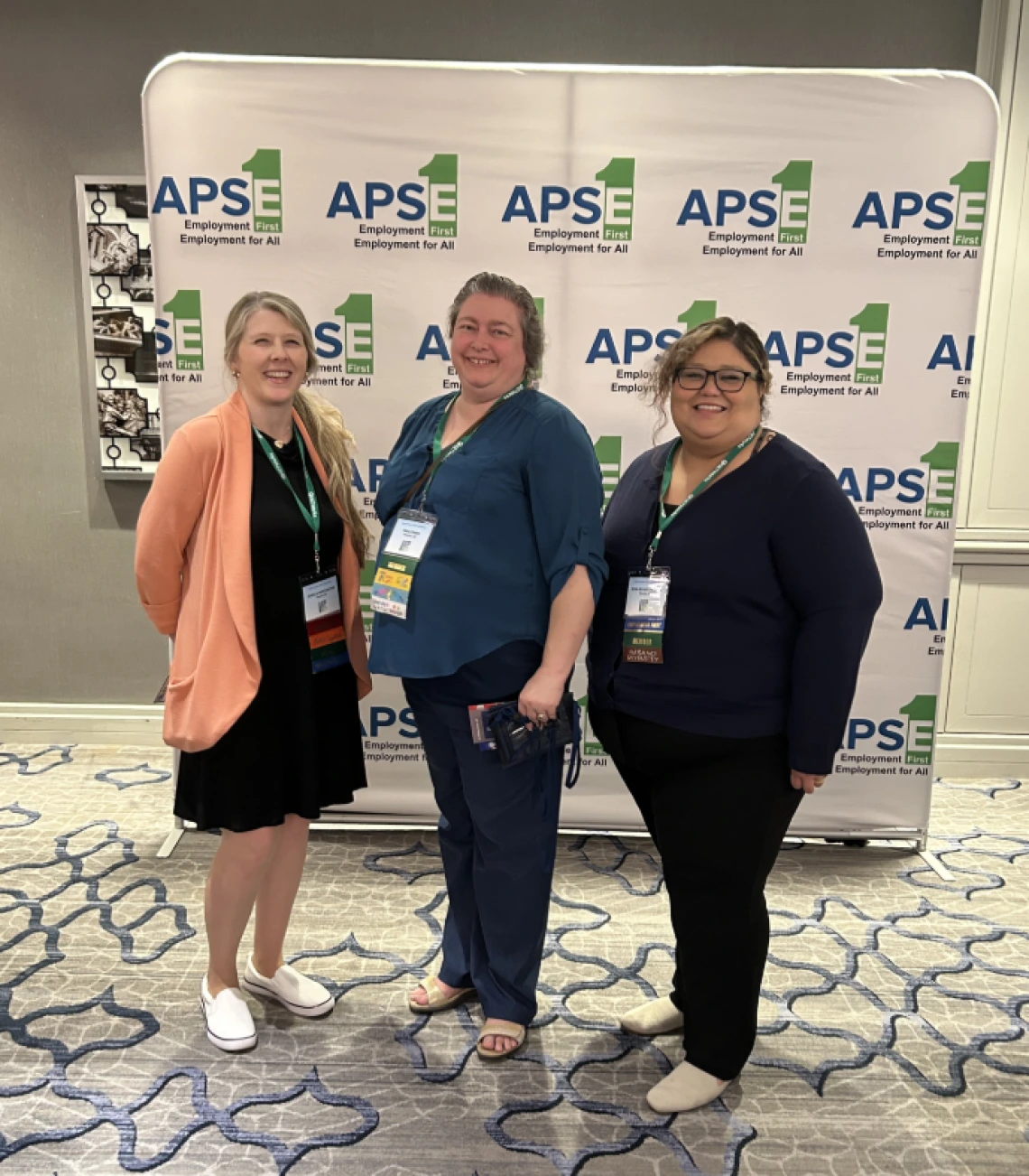 Jessica Winchester (left) and Rosa Rojas Franco (right) pose in front of a banner with the logo of the APSE Employment First, Employment for All Conference. 