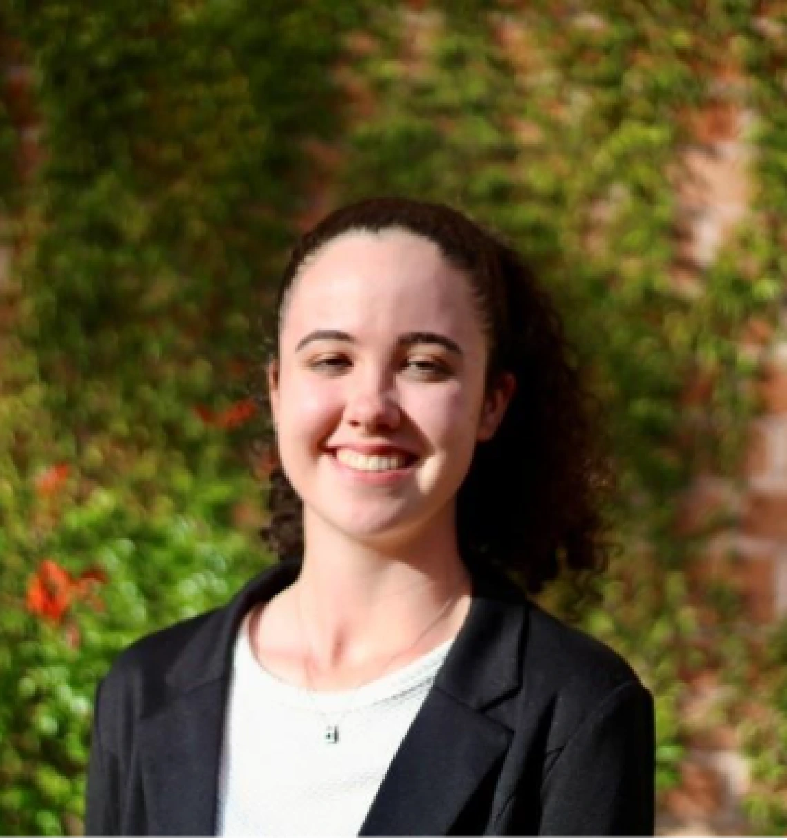 Smiling young woman with curly brown hair pulled back in a ponytail, wearing a black blazer over a white top, standing outdoors in front of a leafy green background