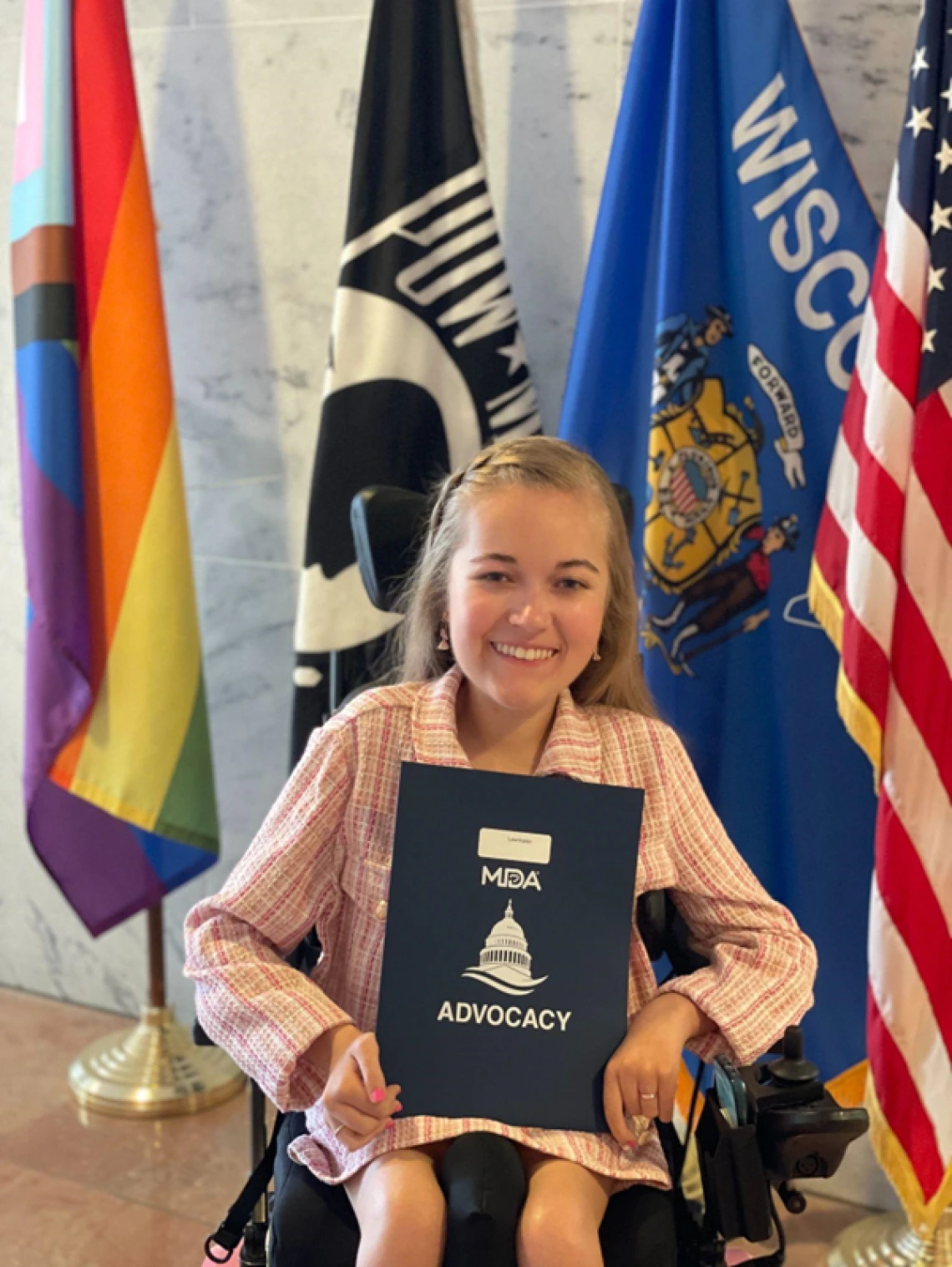 Reagan, a white woman in a pink suit and who sits in a power wheelchair, holds up a folder that says MDA Advocacy on the cover. Reagan sits in front of several inclusive flags, alongside the state of Wisconsin flag and the American flag. Reagan smiles at the camera.