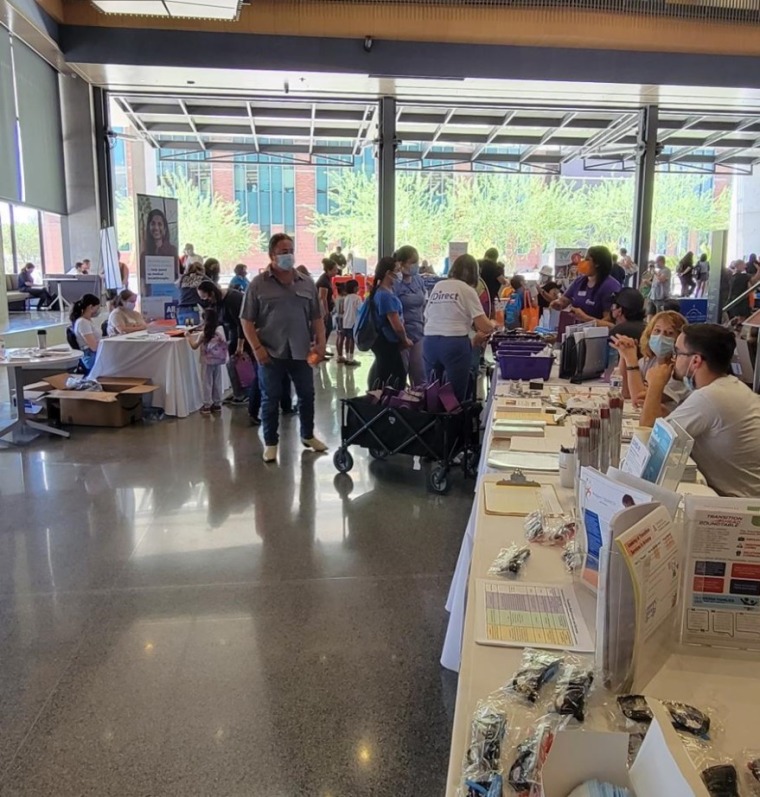 A photograph taken on the floor of the Backpacks & Boosters event. Event attendees are observing and interacting with community partners and resources at several booths.