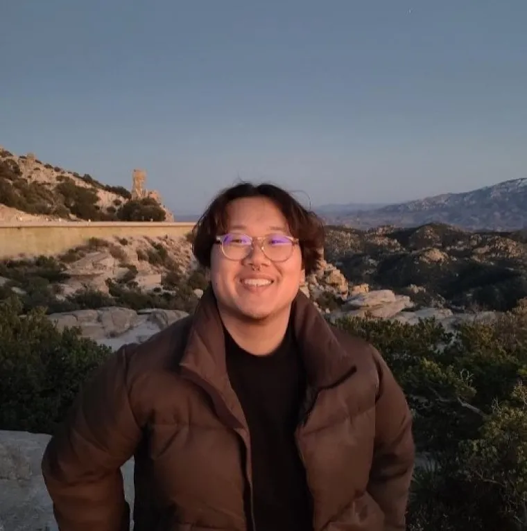 Andrew, a Chinese American man with dark brown hair, is outside in front of a mountain range. He wears clear-rimmed glasses and smiles at the camera. He also wears a black mock-neck and a brown puffer jacket. 