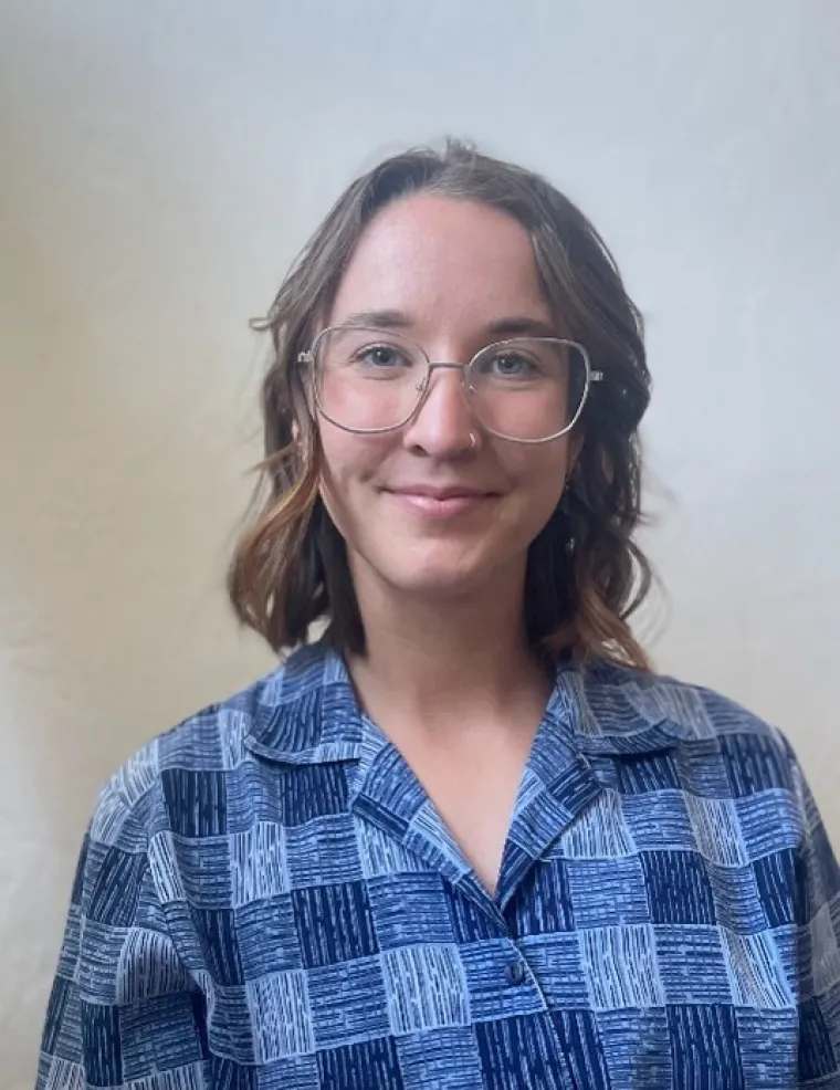 Audrey, a white woman with shoulder length brunette hair, standing in front of a white background. She wears silver rimmed glasses and smiles at the camera. She is also wearing a  blue checkered collared shirt. 