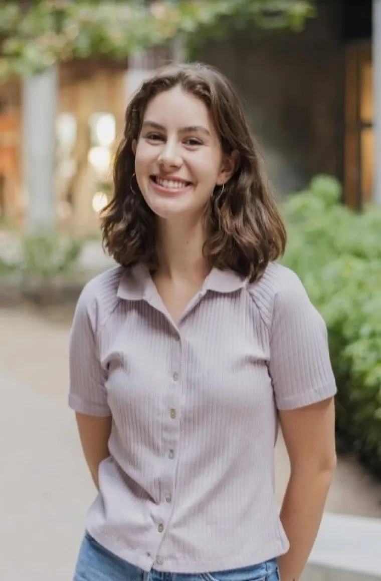 Cate has shouldner length burnete hair and is smiling at the camera, wearing a tan collared button up shirt and blue jeans.