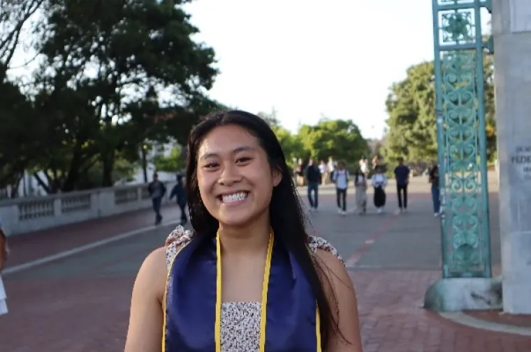 Cosette, an Asian woman with long black hair, smiling at the camera and standing outside in front of a big teal arch gate.  She is wearing a white dress with floral designs with a navy blue graduation stole with gold outlines. 