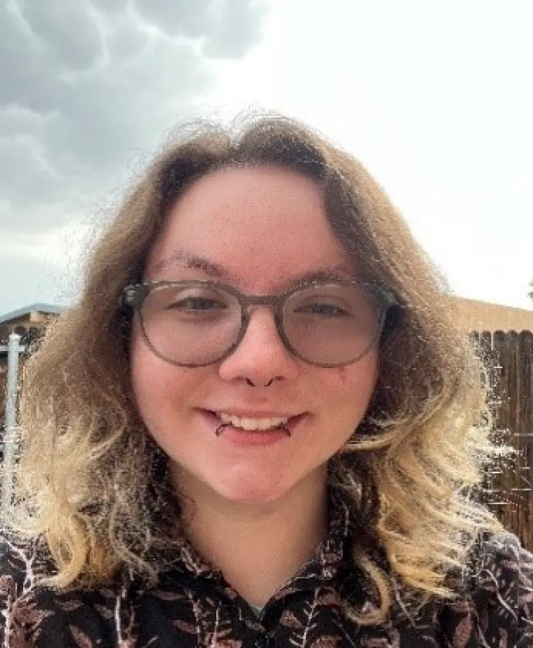 Elexander, a white man with shoulder-length brown hair with the ends bleached, is smiling at the camera. He has round green glasses, a black septum ring and two black lip rings. He is also wearing a black button-up shirt with brown and white leaves on it