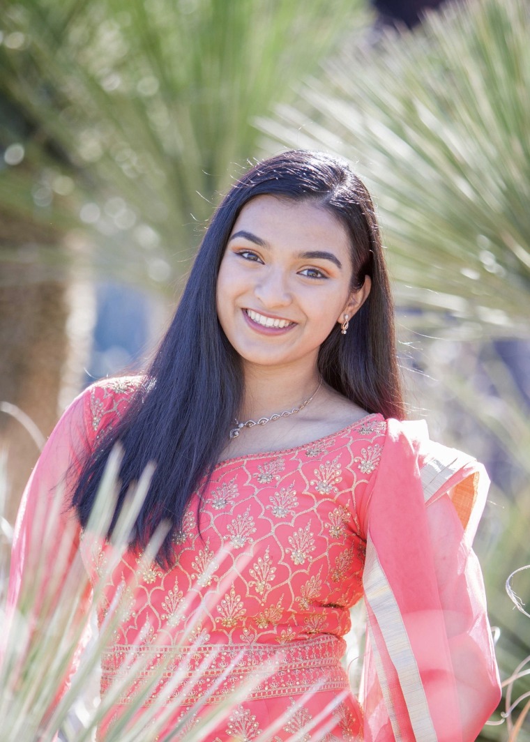 Freya, a woman of color with long black hair, is outside in front of a palm tree. She smiles at the camera and wears a traditional Indian dress in pink with gold detailing. 