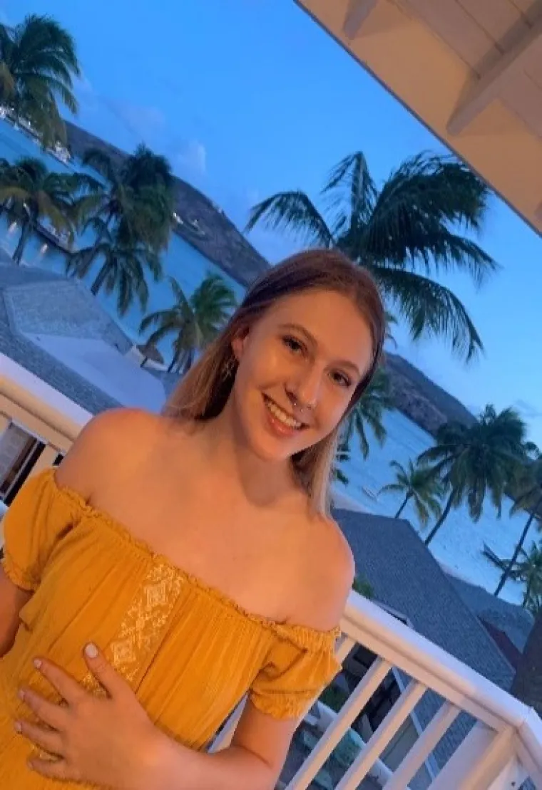 Gabby, a white woman with blonde hair and blue eyes is standing on a balcony in front of a blue backdrop of palm trees and the Caribbean ocean, wearing an off-shoulder yellow sundress.