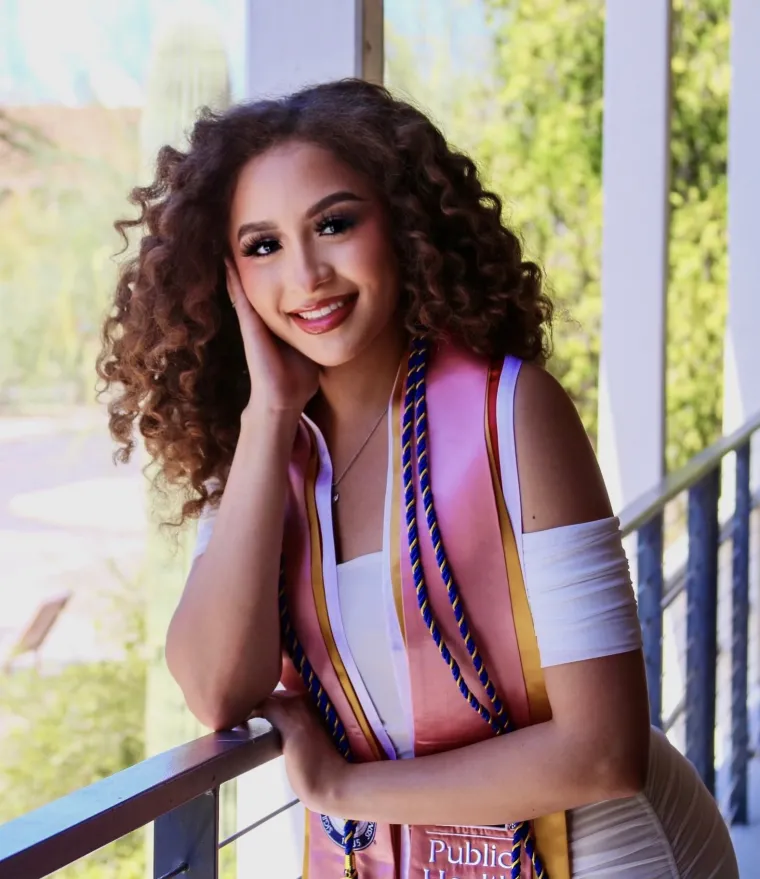 A young multi-ethnic woman with curly brown hair wearing a white dress and graduation stoles with desert landscape in the background.