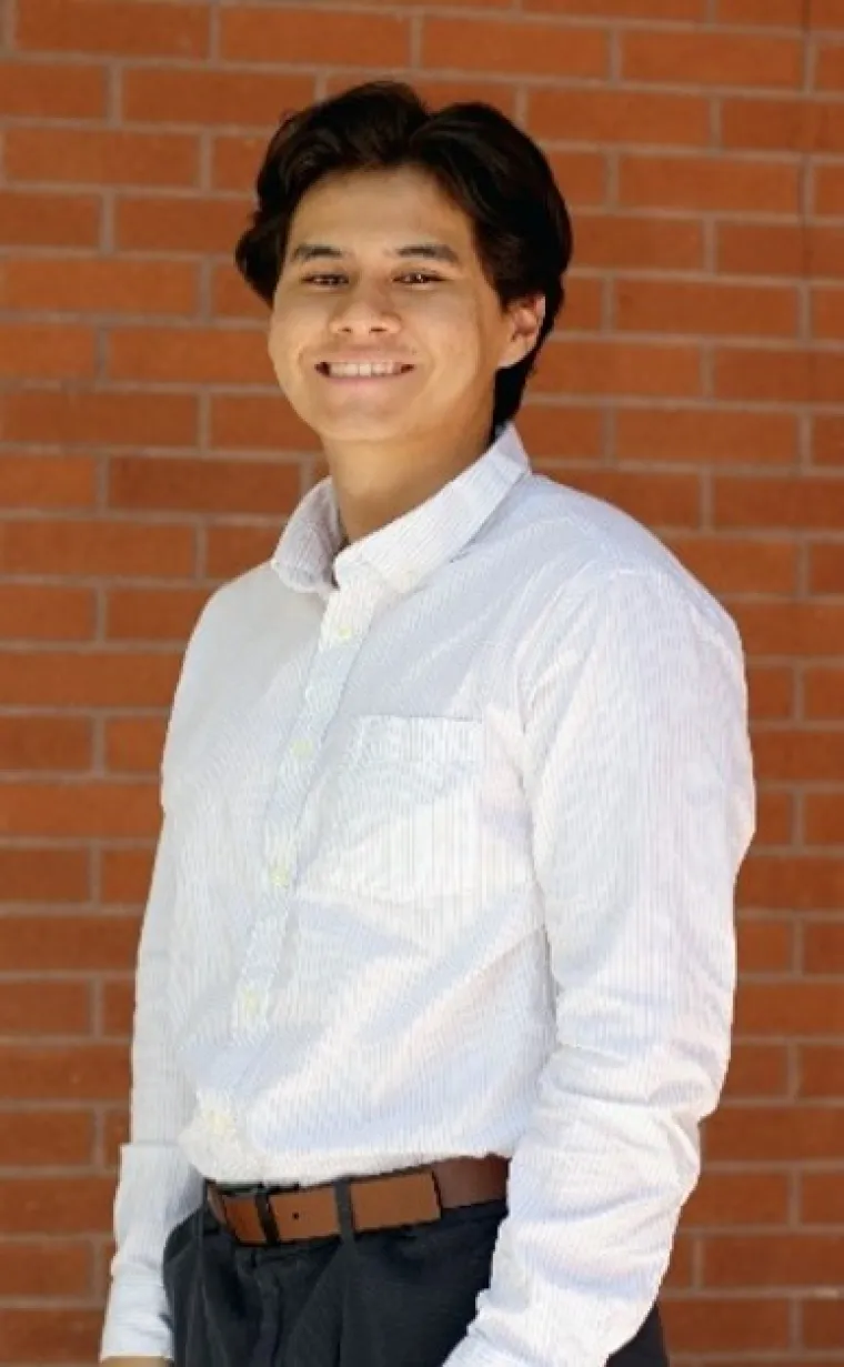 Joseph, a dark-skinned person with long brown hair, is outside on a bright day in front of a red brick wall. He wears a striped white shirt with black pants and a brown belt, and smiles at the camera.