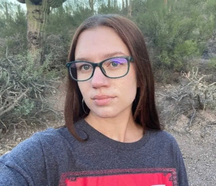 Kirsten, a white woman with red hair is outside. She is wearing green glass and a red and gray t-shit, standing in front of a desert landscape. 