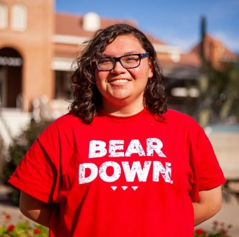 Lexicon, is facing the camera and smiling. His hands are behind his back and he stands in front of Old Main, which is blurred in the background. He is wearing a red shirt that says "BEAR DOWN" in all caps, and his hair is shoulder-length and curly. 