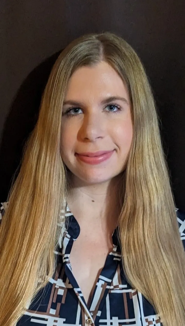 A white woman with blonde hair is smiling in front of a brown background. She is wearing a navy blouse that has white and copper geometric patterns. 